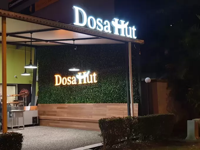 The Dosa Hut Aspley shopfront at night, its lit Dosa Hut signage above a green hedge wall, timber bench seating and a covered outdoor terrace